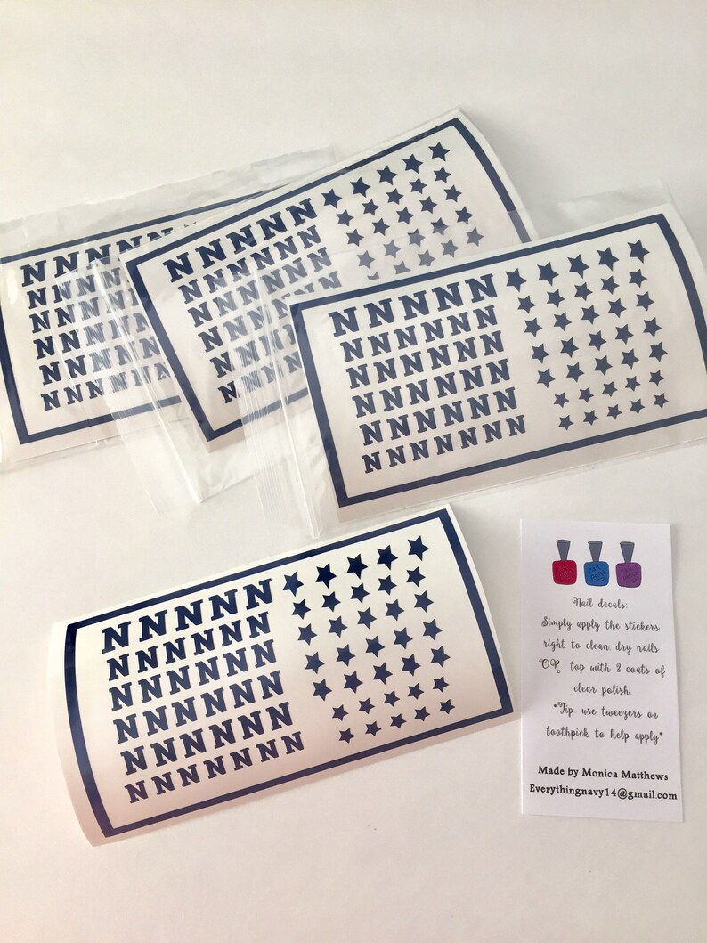May include: Four rectangular sheets of dark blue nail decals featuring a repeating pattern of the letter N and five-pointed stars, enclosed in clear plastic sleeves. A small white instruction card with three nail polish bottle icons is included.