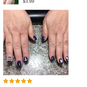 May include: A set of navy blue nail decals applied to fingernails, featuring white designs including a letter N, a nautical anchor, a five-pointed star, and horizontal stripes. The decals are applied over a solid dark blue nail polish base.