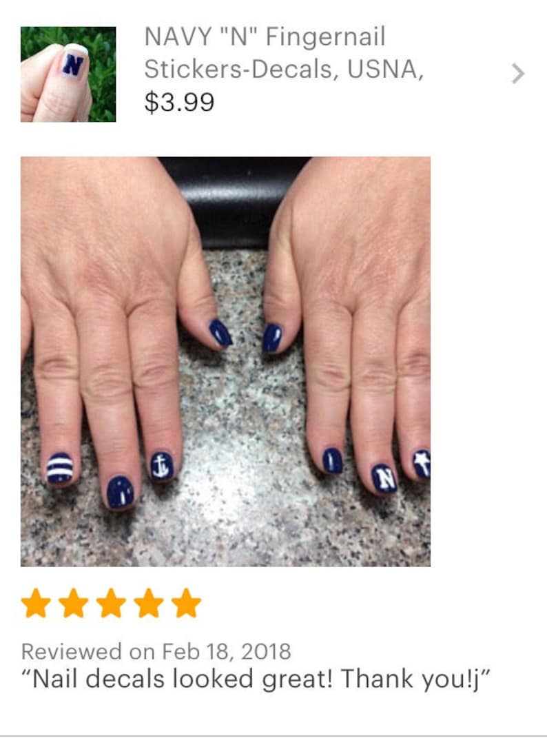 May include: A set of navy blue nail decals applied to fingernails, featuring white designs including a letter N, a nautical anchor, a five-pointed star, and horizontal stripes. The decals are applied over a solid dark blue nail polish base.