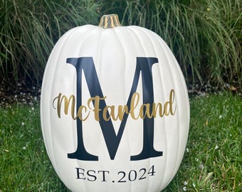 May include: A white artificial pumpkin with a gold-colored stem. The front features a large dark navy blue letter M, the name McFarland in gold script lettering overlaid across the center, and the text EST. 2024 in dark navy blue at the bottom.