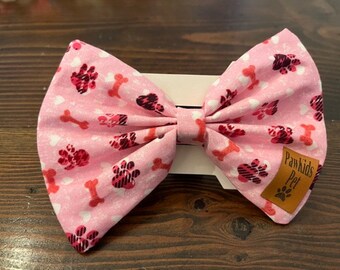 May include: Pink bow tie with a paw print and bone pattern. The bow tie has a brown tag that says "Pawkids Pet".
