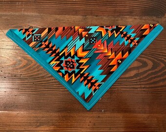 May include: A turquoise blue bandana with a geometric pattern in shades of orange, brown, and black. The bandana is folded into a triangle shape.