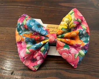 May include: A large floral bow with a variety of colors, including pink, blue, yellow, and green. The bow is made of fabric with a watercolor-like floral print. The bow is tied with a white ribbon and has a tag that says "handmade with love".
