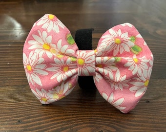 May include: A pink bow tie with a white daisy print. The bow tie is made of fabric and has a black elastic band.