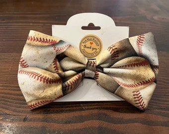May include: A brown and white bow tie with a baseball print. The bow tie is on a white card with a brown circle that says "Hand Made With Love" and "Thank You".