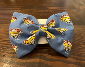 May include: A blue bow tie with a repeated pattern of the Superman logo in yellow, red, and white. The logo includes the trademark symbol "TM".