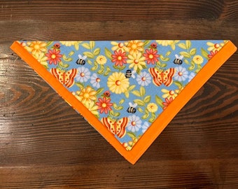 May include: A blue bandana with a floral pattern featuring butterflies, bees, and flowers. The bandana has an orange border.