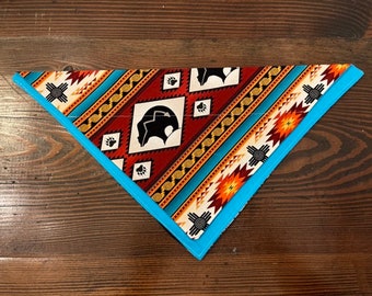 May include: A triangular dog bandana with a blue border and a colorful geometric pattern featuring a bear silhouette and Native American symbols.
