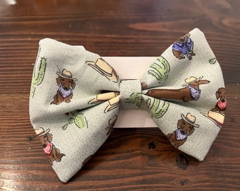 May include: A light blue bow tie with a pattern of brown dachshunds wearing cowboy hats and bandanas. The dachshunds are surrounded by cacti and cowboy hats.