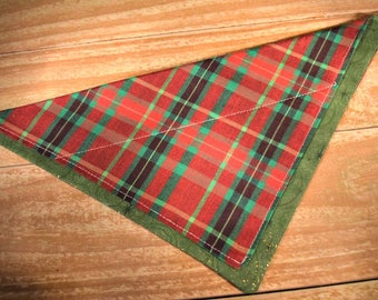 May include: A red, green, and black plaid bandana with a green border. The bandana is folded into a triangle shape and is lying on a wooden surface.