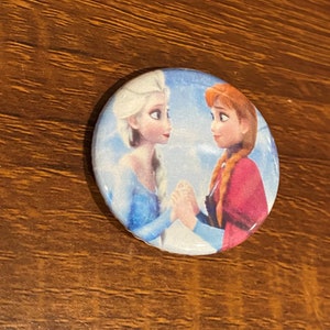 May include: A round button featuring a colorful image of two animated characters, Elsa and Anna from the movie Frozen, standing side by side with their hands clasped together. The background is a light blue with a white sparkle effect.