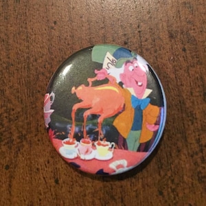 May include: A round button featuring a cartoon image of the Mad Hatter from Alice in Wonderland pouring tea from a teapot shaped like a flamingo. The Mad Hatter is wearing a green hat, a yellow jacket, and a blue bow tie. The background is a dark green and black pattern.