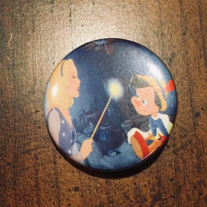 May include: A round pin featuring a cartoon image of a fairy with blonde hair and a blue dress holding a wand, and a boy with brown hair wearing a blue and white shirt and a red hat. The background is a dark blue with stars.