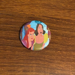 May include: A round button featuring a cartoon image of two women with red and blonde hair. The woman with blonde hair is wearing a blue and yellow dress and the woman with red hair is wearing a purple dress. The women are looking at each other and appear to be arguing.
