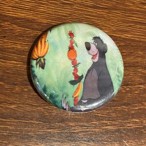 May include: A round button featuring a cartoon image of Baloo the bear from Disney's The Jungle Book. Baloo is wearing a green shirt and holding a stick with fruit on it. The background is a green and brown jungle scene.