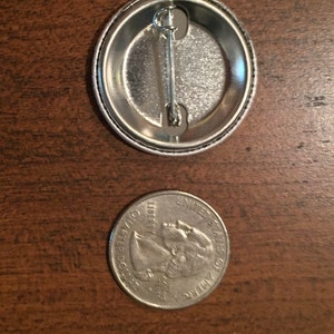 May include: The back of a silver button with a safety pin attached. The button is about the size of a US quarter.