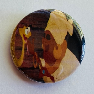 May include: A round button with a cartoon image of a brown-skinned character wearing a white turban and a yellow snake with a red tongue. The background is a dark brown wood.