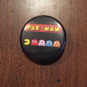 May include: A black button with a yellow Pac-Man and four ghosts in red, pink, blue, and orange. The button says "PAC-MAN" in yellow letters.
