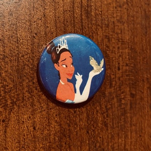 May include: A round button featuring a cartoon image of a princess with a crown, wearing a blue dress and holding a small green frog. The background is a dark blue with white stars.