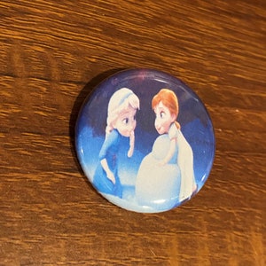 May include: A round button with a picture of two young girls from the movie Frozen. The girls are standing in front of a blue and white background. The girl on the left is wearing a blue dress and has long blonde hair. The girl on the right is wearing a white dress and has long red hair.