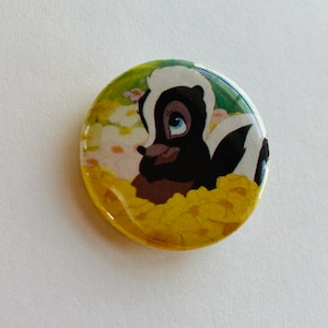 May include: A round button featuring a cartoon image of a skunk with black and white fur, surrounded by yellow flowers. The skunk has a pink nose and blue eyes.