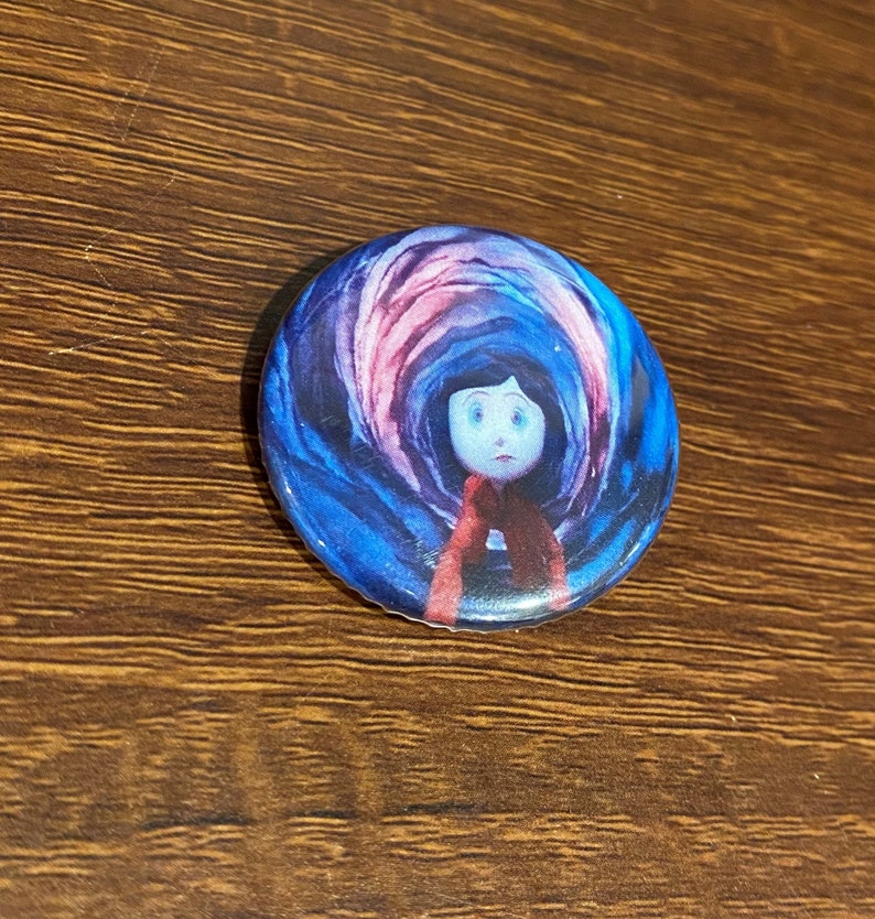 May include: A round button with a colorful image of a girl with dark hair and a red scarf standing in front of a swirling blue and pink background.