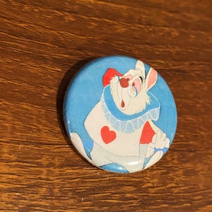 May include: A round button featuring a cartoon image of the White Rabbit from Alice in Wonderland. He is wearing a blue and red waistcoat with a white shirt and a red heart on his chest. The background is blue.