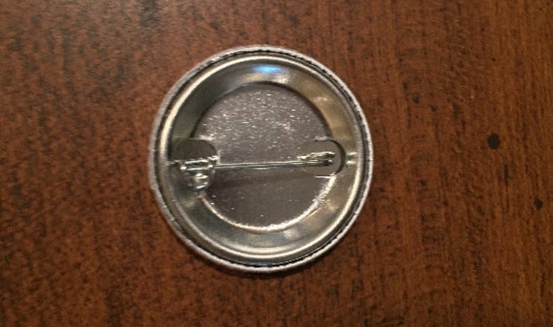 May include: The back of a round, silver button with a pin attached. The button has a white border.