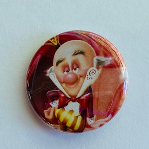 May include: A round button featuring a cartoon character with a bald head, a red bow tie, and a purple and gold outfit. The button has a glossy finish and a red border. The character has a large nose and is smiling.