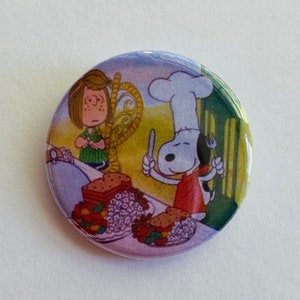 May include: A round button featuring Snoopy in a chef's hat holding a fork and knife, with a girl looking on. The image depicts a meal with plates of food. The button has a glossy finish and a colorful design.