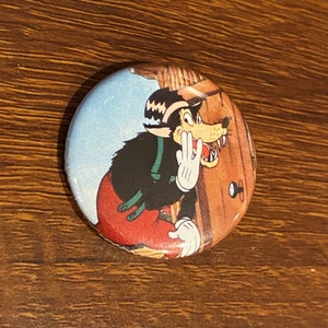 May include: A vintage button featuring a cartoon wolf with a red and black outfit, peeking around a wooden door.
