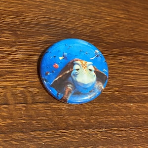 May include: A blue and green button with a cartoon turtle from the movie Finding Nemo. The turtle is smiling and has a blue background with other fish swimming around it.