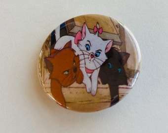 May include: A round button featuring a cartoon image of three cats. The cats are a white cat with a pink bow, a brown cat, and a black cat. The cats are sitting on a wooden surface.