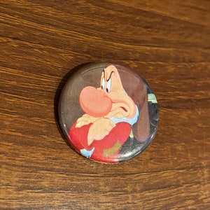 May include: A round button with a cartoon image of a grumpy dwarf with a large red nose. The dwarf is wearing a blue and brown hat and a red shirt.