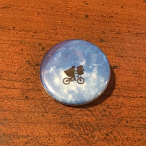 May include: A blue and white button with a silhouette of a bicycle and two figures flying in the sky. The figures are riding the bicycle with the front wheel off the ground.