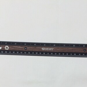 Puede incluir: Una regla negra y marr&oacute;n con la palabra "WESTCOTT" impresa en ella. La regla mide 30,5 cm de largo y tiene marcas para cada cent&iacute;metro y cada 1/16 de pulgada. La regla est&aacute; hecha de metal y tiene un acabado de madera.