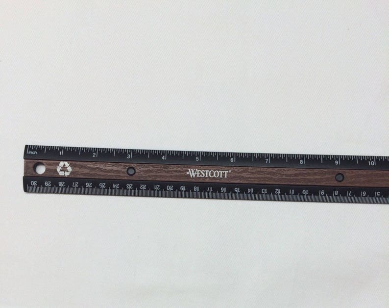 Puede incluir: Una regla negra y marr&oacute;n con la palabra "WESTCOTT" impresa en ella. La regla mide 30,5 cm de largo y tiene marcas para cada cent&iacute;metro y cada 1/16 de pulgada. La regla est&aacute; hecha de metal y tiene un acabado de madera.