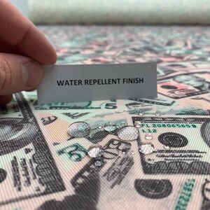 Pode incluir: Tecido repelente &agrave; &aacute;gua com uma estampa de notas de d&oacute;lar americano. O tecido &eacute; mostrado com gotas de &aacute;gua sobre ele, demonstrando suas propriedades repelentes &agrave; &aacute;gua. O texto "WATER REPELLENT FINISH" &eacute; vis&iacute;vel em um cart&atilde;o branco.