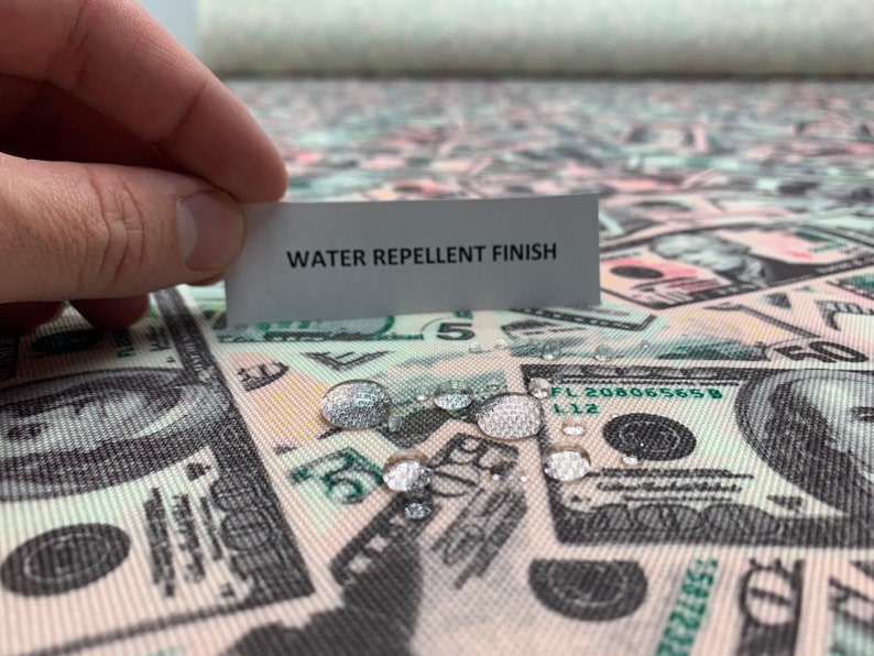 Pode incluir: Tecido repelente &agrave; &aacute;gua com uma estampa de notas de d&oacute;lar americano. O tecido &eacute; mostrado com gotas de &aacute;gua sobre ele, demonstrando suas propriedades repelentes &agrave; &aacute;gua. O texto "WATER REPELLENT FINISH" &eacute; vis&iacute;vel em um cart&atilde;o branco.