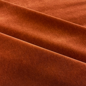 May include: Close-up of a rich, rust-colored fabric with a soft, velvety texture. The fabric is folded in a smooth, elegant drape, highlighting its luxurious feel.