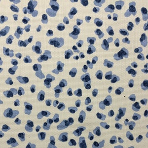 May include: Close-up of a cream-colored fabric with a repeating pattern of blue, irregular shapes. The shapes vary in size and shade, creating a spotted or speckled effect. The fabric has a woven texture.