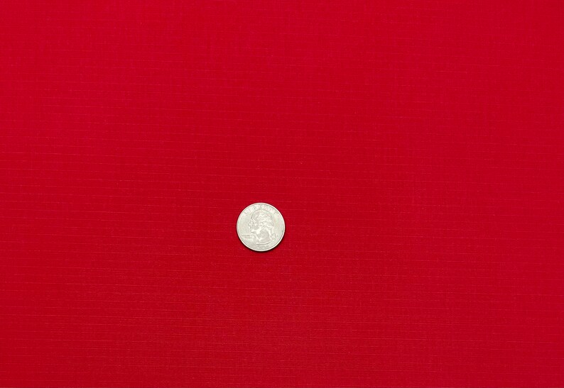 May include: A close-up of a solid red fabric with a subtle textured pattern. A silver coin is placed in the center of the fabric.