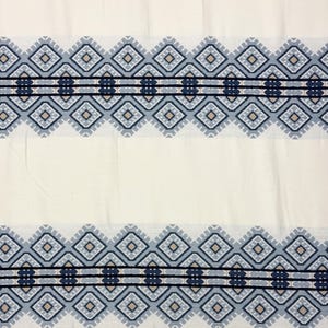 Peut inclure: Tissu crème avec deux bandes de motifs géométriques dans des tons de bleu, beige et noir. Le motif présente des formes de losanges et des lignes horizontales, créant une bordure symétrique. Le tissu semble être une matière tissée.