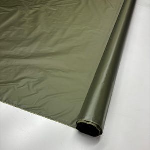 Puede incluir: Rollo de tela verde oliva. La tela tiene una superficie lisa y ligeramente brillante, con pliegues y arrugas visibles. El rollo está parcialmente desenrollado, revelando la textura y el color de la tela. La tela parece ser de peso medio.