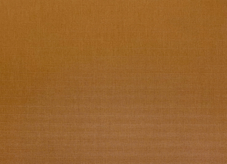 May include: A close-up of a textured, golden-brown fabric. The fabric has a subtle grid pattern created by fine, horizontal and vertical lines. The overall appearance is a solid, warm colour with a slightly rough texture.