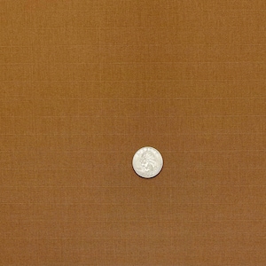 May include: A silver coin, a quarter, sits on a brown, textured fabric. The fabric has a grid pattern, with horizontal and vertical lines creating a subtle woven look. The coin shows a profile.
