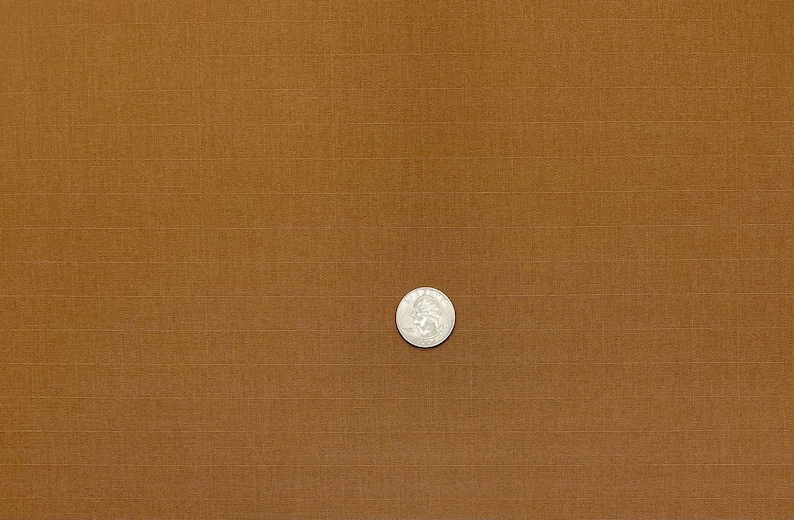 May include: A silver coin, a quarter, sits on a brown, textured fabric. The fabric has a grid pattern, with horizontal and vertical lines creating a subtle woven look. The coin shows a profile.