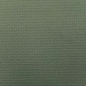 May include: Close-up of a textured fabric in a muted green color. The fabric has a subtle, grid-like pattern, creating a slightly raised, tactile surface. The overall appearance is uniform, with a consistent color and texture across the frame.