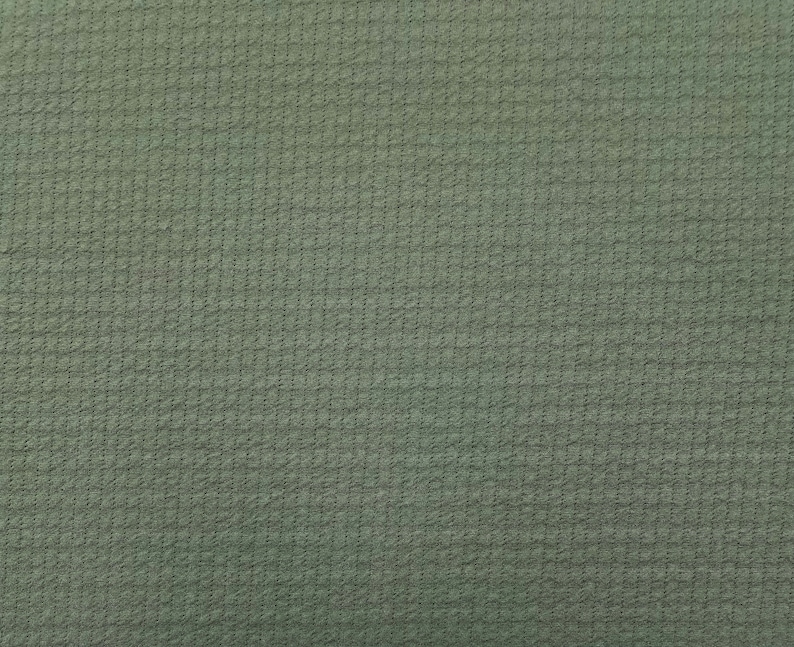 May include: Close-up of a textured fabric in a muted green color. The fabric has a subtle, grid-like pattern, creating a slightly raised, tactile surface. The overall appearance is uniform, with a consistent color and texture across the frame.