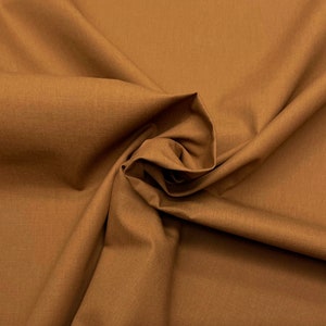May include: Close-up of a fabric swatch in a warm, earthy brown hue. The material is tightly woven, showing texture and folds that create shadows. The fabric appears to be a medium-weight textile, suitable for various sewing projects, with a slight sheen.